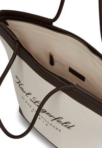 Suuri olkalaukku, jossa on vaaleancreme-värinen kangaspinta, tummanruskeat nahkaosuudet ja vetoketjullinen sisätasku; edessä kohokuvioitu logo.