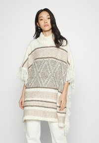 Poncho de malha com gola alta, apresentando riscas horizontais alternadas em bege e branco, padrões de diamante e franjas nos mangas.