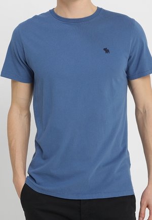Mand iført en ensfarvet blå T-shirt med korte ærmer med en lille mørk logo på venstre bryst og sorte bukser, stående med hænderne i lommerne.