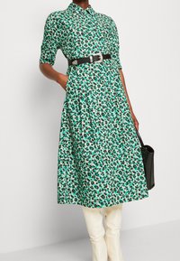 Zielono-biała sukienka w kwiatowy wzór z kołnierzykiem, krótkimi rękawami, czarnym paskiem i kieszeniami. Nosi się ją z jasnymi, dopasowanymi spodniami.