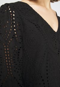 Mustaa brodeerattua kangasta, jossa on V-pääntie, ja jossa on monimutkaisia leikattuja kuvioita sekä tekstuurisia yksityiskohtia hihoissa.