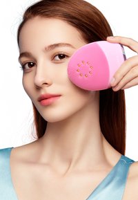 Dispositif de nettoyage facial en silicone rose avec une surface texturée surélevée. L'utilisateur le tient contre sa joue, avec de petits accents dorés.