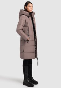 Pikowana zimowa kurtka w jasnym odcieniu mauve, z dużym kapturem, bocznymi zamkami i smukłą sylwetką. W zestawieniu z czarnymi kozakami za kolano.
