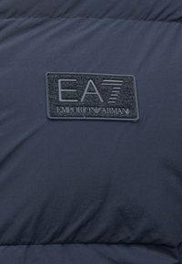 Jaka ar jūras spēku puferiem ar strukturētu melnu auduma plāksteri, uz kura ir iedobts "EA7 Emporio Armani" logo pelēkā krāsā. Gluda virsma.