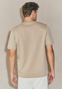 Camiseta de manga corta beige con cuello redondo y textura suave, que presenta un corte holgado. Sin patrones ni acentos visibles.