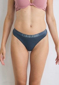 Modell som har på seg en rosa BH og mørkeblå Calvin Klein bikinioverdel, står mot en lys bakgrunn med armene avslappet på sidene.