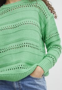 Pull en maille vert clair avec des rayures horizontales et verticales, des détails en tricot ajouré et des poignets côtelés. Porté avec un jean bleu.