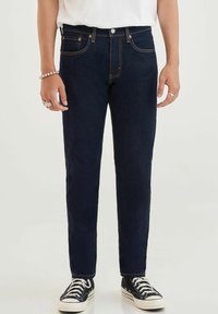 Jeans de mezclilla azul oscuro con corte recto, diseño de cinco bolsillos, costuras en contraste y cierre de botón, combinados con zapatillas negras.