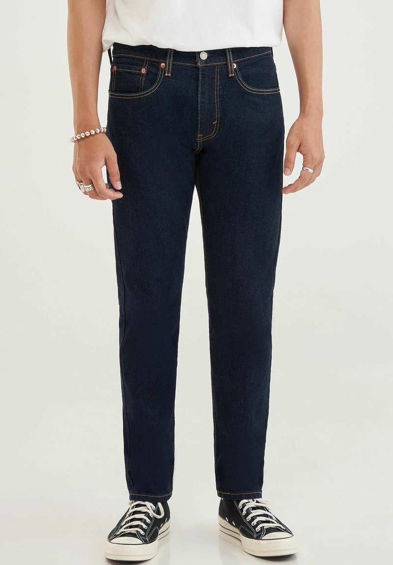 Jeans de mezclilla azul oscuro con corte recto, diseño de cinco bolsillos, costuras en contraste y cierre de botón, combinados con zapatillas negras.