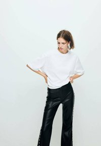 T-shirt blanc oversize à manches courtes, associé à un pantalon noir en simili cuir taille haute avec des œillets latéraux.