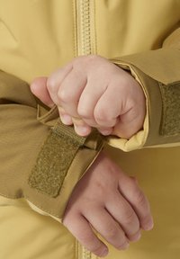 Geltona medžiaginė striukė su tekstūruotu paviršiumi, turinti priekinį užtrauktuką. Rankovė su reguliuojamu Velcro užsegimu, atidengiančiu minkštą audinio pamušalą.