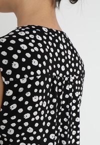 Černý top s bílým tečkovaným vzorem. Tkanina má texturovaný pocit, s plisovaným detailem na zádech pro větší objem.