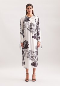 Langes, weißes Kleid mit schwarzen architektonischen Skizzen, gesammeltem Ausschnitt und langen Ärmeln mit Kordelzugdetails. Getragen mit schwarzen Knöchelriemen.