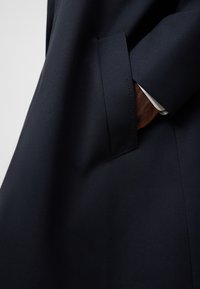 Cappotto navy su misura in tessuto testurizzato, con tasca angolata e logo in rilievo discreto, e una fodera interna a contrasto visibile al polsino.