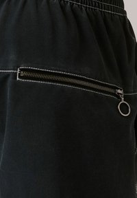 Fekete szövet joggerek, elasztikus derékkal és cipzáras zsebbel, ezüst színű fémrészekkel és kontrasztos varrással. Strukturált felület.