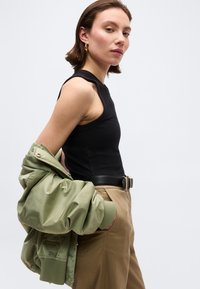 Γυναίκα με μαύρο τοπ χωρίς μανίκια και μπεζ παντελόνι κρατά ανοιχτό πράσινο σακάκι στον ώμο της μπροστά σε απλό φόντο.