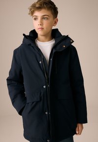 Parka blu navy con cappuccio, chiusura con cerniera, due tasche frontali e bottoni automatici; realizzata in tessuto liscio e resistente.