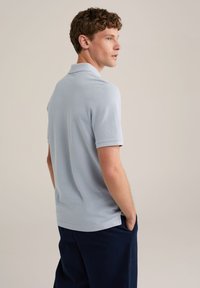 Tricou polo albastru deschis cu guler clasic, mâneci scurte și un material texturat. Tricoul are o croială lejera și slituri pe laterale la tiv.