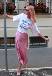 Woman in sunglasses holds a pole, wearing a white "Pimp my shrimp" t-shirt and pink skirt with green spots, standing on a city sidewalk.