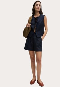 Crop top en lin marine avec un nœud à l'avant, associé à un short assorti. Dispose d'un design sans col et de poches latérales. Des chaussures plates marron complètent le look.