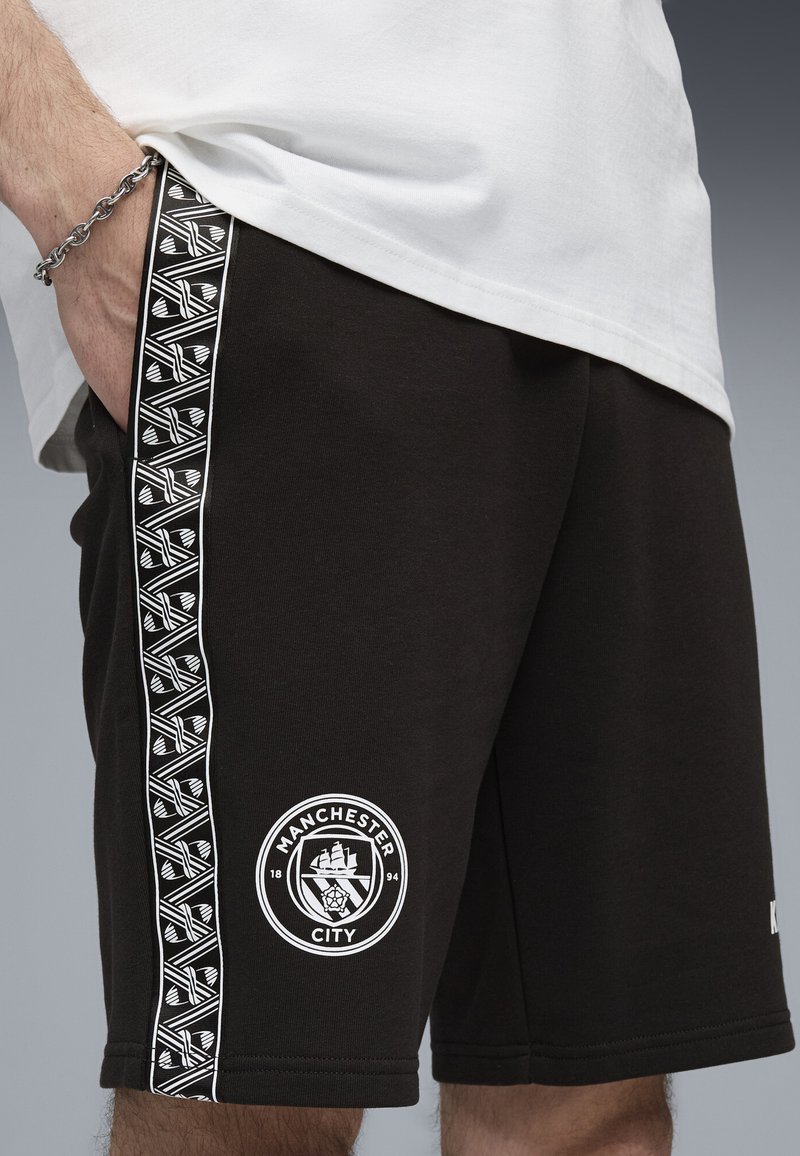 Svarta shorts i bomull med en vit logotyp som föreställer ett skepp och en ros, accentuerade av en mönstrad sidostråk.