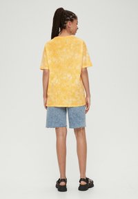 Żółta koszulka tie-dye z krótkimi rękawami, zestawiona z wyblakłymi dżinsowymi szortami. Model nosi czarne sandały i duże okrągłe kolczyki.