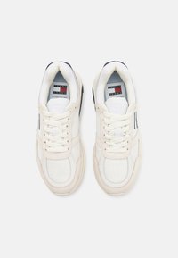 Zapatillas blancas y crema con parte superior de tela texturizada, suela de goma y acentos en azul marino. Presentan cordones atados y una etiqueta de marca en la lengüeta.