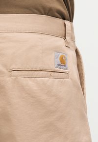 Τσέπη μπεζ παντελονιού Carhartt με λογότυπο, συνδυασμένη με καφέ πουκάμισο, κοντινή λήψη που δείχνει την υφή του υφάσματος και τις λεπτομέρειες των ραφών.