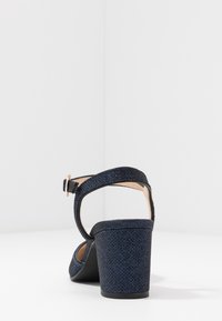 Mörkblå häl sandalett med en texturerad ovandel, ankelrem och en tjock klack. Har ett spänne och en slät foder på insidan.