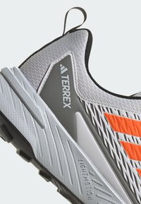 Lichtgewicht grijze sportschoen met textuur bovenkant, oranje accenten en een grijs paneel met het "Terrex" logo; rubberen zool met grip.