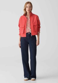 Giacca rossa corta con due tasche frontali, abbinata a una camicia a righe e pantaloni larghi in denim scuro. Sneakers chiare completano il look.