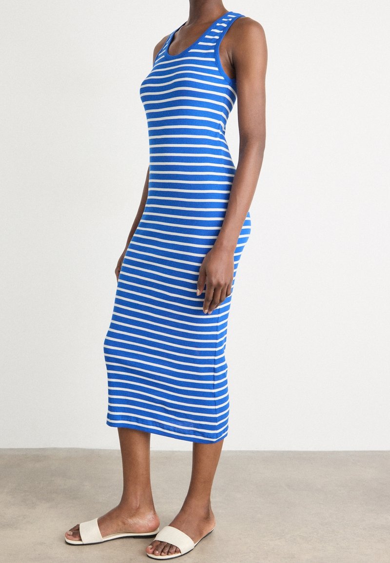 Femme portant une robe midi sans manches à rayures horizontales bleues et blanches et des sandales blanches, debout sur un sol gris.