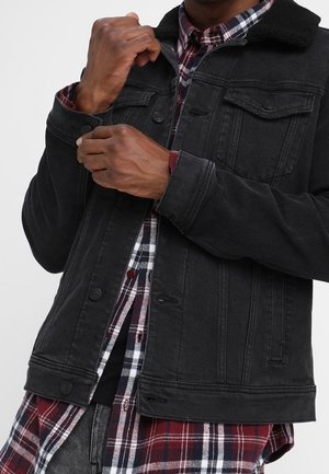 Mężczyzna poprawiający mankiet czarnej kurtki dżinsowej noszonej na czerwono-czarnej koszuli w kratę.