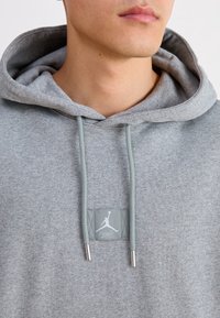 Grå sweatshirt med huva, med en framstående patch-logga och ljusgrå dragsnören med metalländar. Mjuk, texturerad tyg.