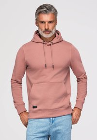 Mann mit grauen Haaren und Bart, der einen staubrosa Hoodie und hellblaue Jeans trägt, steht vor einem schlichten weißen Hintergrund.