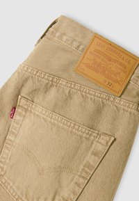 Μπεζ τζιν Levi's διπλωμένα ώστε να φαίνεται η πίσω τσέπη, η κόκκινη ετικέτα Levi's και η ταμπέλα από μαύρο δέρμα Levi Strauss & Co. με τη σήμανση μεγέθους W32L.