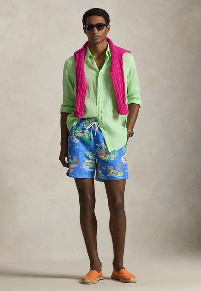 Světlá zelená lněná košile, růžový pletený svetr přehozený přes ramena, modré plavky s barevným tropickým potiskem, oranžové espadrilky, sluneční brýle.