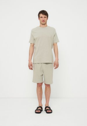 Άνδρας που στέκεται σε λευκό φόντο με ανοιχτό μπεζ κοντομάνικο πουκάμισο, αντίστοιχα σορτς και μαύρα σανδάλια.