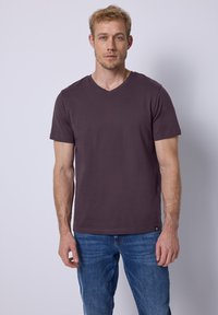 Camiseta de algodón de cuello en V y manga corta en púrpura oscuro, combinada con jeans azules, exhibida en un modelo masculino sobre un fondo claro.