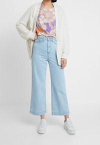 Cárdigan crema de punto sobre una blusa estampada en púrpura y naranja, combinado con unos vaqueros de pierna ancha de tiro alto en azul claro y zapatillas blancas.