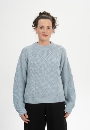Femme aux cheveux bouclés portant un pull bleu clair en maille avec des motifs torsadés et un pantalon noir, debout contre un fond blanc.