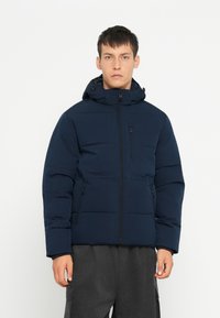 Marineblå polsteret jakke med hætte, lynlåse i siderne og en brystlomme. Har en struktureret overflade og et afslappet fit design.