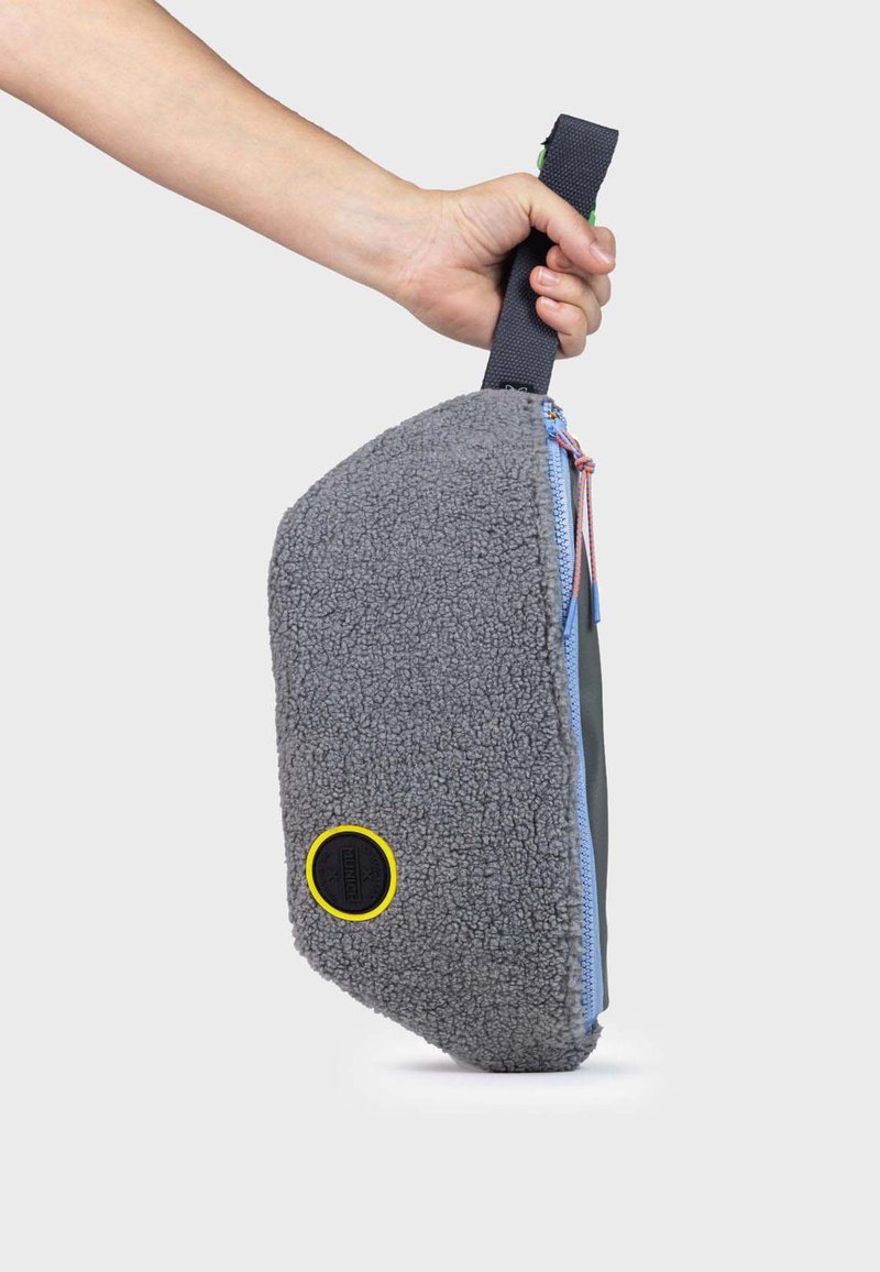 Bolsa de tela gris con un exterior de forro polar texturizado y cremallera azul, que presenta un acento circular amarillo y un asa negra robusta.