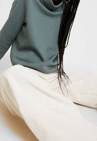 En person sitter klädd i en tealfärgad sweatshirt och vita kordbyxor, med långt flätat hår hängande åt sidan.