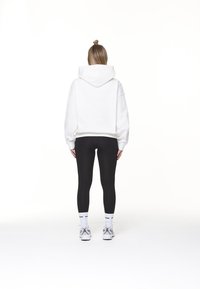 Sudadera blanca recortada con capucha, combinada con leggings negros. El modelo lleva zapatillas blancas y calcetines con letras negras, visto desde atrás.