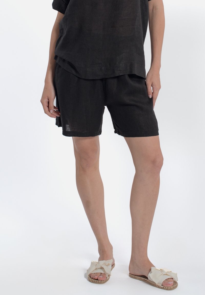 Pantaloni corti neri in lino abbinati a una camicia nera in lino dal taglio morbido, completati da sandali chiari con dettagli annodati sulla parte anteriore.