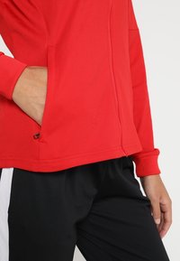 Rode sporttop met lange mouwen, voorzien van een zijzak en een gladde, platte textuur. Gecombineerd met zwarte broek met witte accenten.