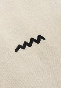 Beżowa tkanina z teksturowanym czarnym wzorem w zygzaki na powierzchni. Projekt jest minimalistyczny i wyraźnie wyśrodkowany.