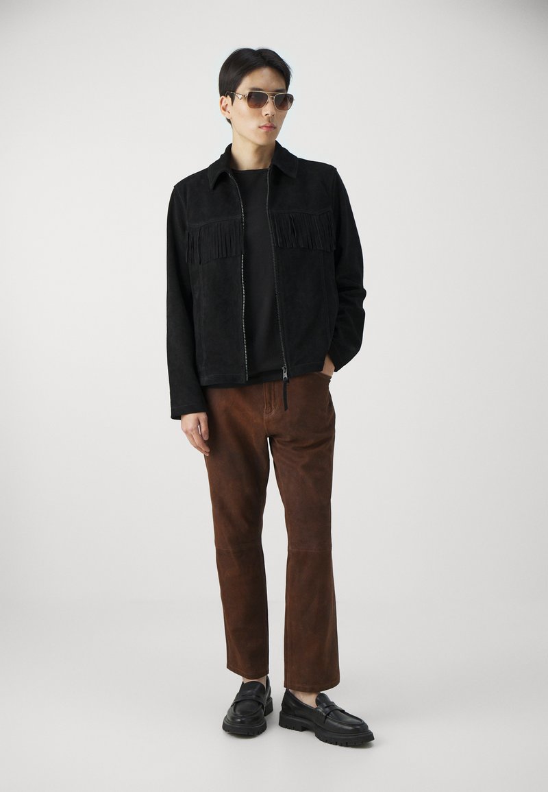 Homme portant une veste à franges noire, une chemise noire, un pantalon en daim marron, des mocassins noirs et des lunettes de soleil, se tenant devant un fond clair.