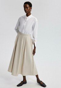 Witte blouse met lange mouwen en een kraag, gecombineerd met een beige A-lijn rok met een zijkant rij knopen en een gladde textuur. Zwarte schoenen.